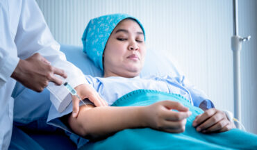 A imagem contém uma mulher com um lenço na cabeça, deitada em uma maca, tomando medicamento.