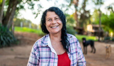 Imagem de uma mulher sorrindo, em representação à saúde da mulher, e ao fundo uma paisagem de campos verdes, árvores, casa e cachorros.