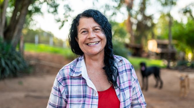 Imagem De Uma Mulher Sorrindo, Em Representação à Saúde Da Mulher, E Ao Fundo Uma Paisagem De Campos Verdes, árvores, Casa E Cachorros.