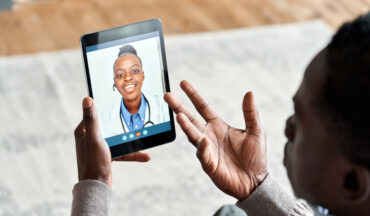 Imagem de um homem em casa segurando um tablet nas mãos, em consulta online com uma médica.