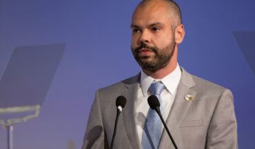 Imagem de uma fotografia do prefeito Bruno Covas, acometido pelo Câncer de Cárdia. Bruno está de terno e gravata, e à frente dois microfones