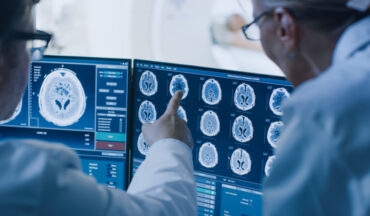 Imagem de dois médicos de costas, analisando um quadro clínico de um paciente, por meio de radiografias do cérebro