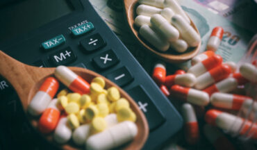 imagem de pílulas medicativas em uma superfície plana, alguns aglomeradas em duas colheres de madeira que estão próximas à uma calculadora em representação à isenção de impostos aos medicamentos que tratam o câncer