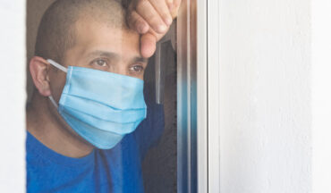 imagem de um homem careca e usando máscara olhando o ambiente externo através de uma janela